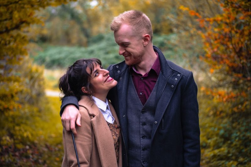 Couple enlacé au milieu des feuillages d'automne, échangeant un regard et un grand sourire