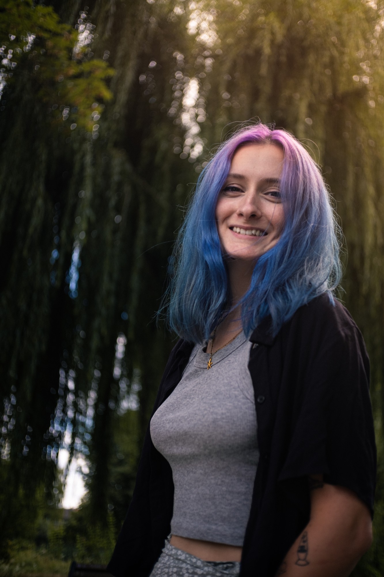 Portrait souriant en extérieur, cheveux bleus et roses, sous les branches d'un saule pleureur