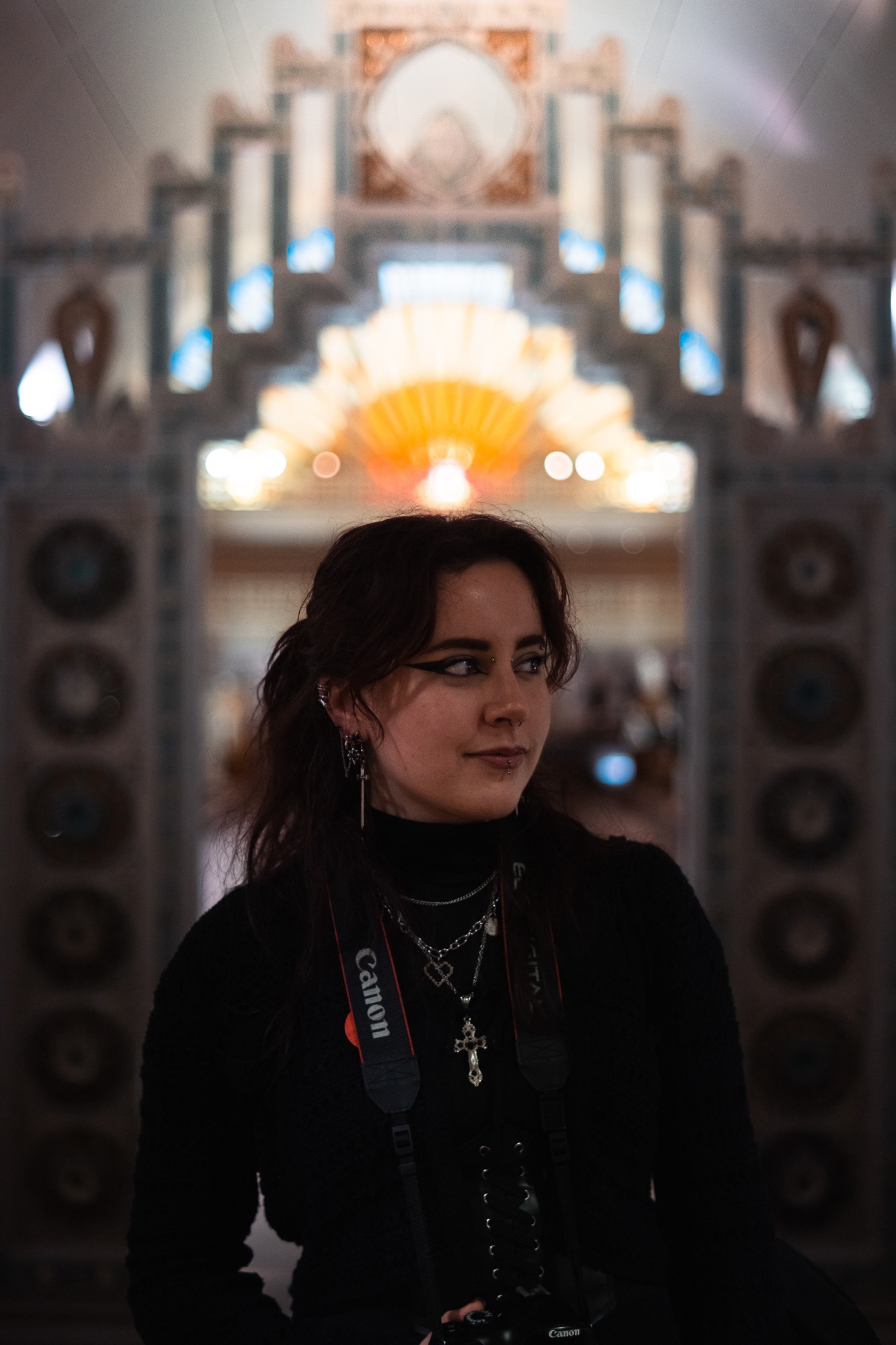 Portrait d'une jeune femme aux cheveux bruns, appareil photo Canon en bandoulière, devant un décor Art déco lumineux et flou